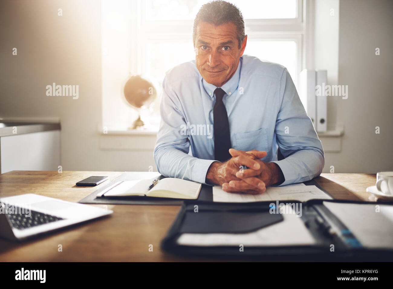 Smiling mature businessman wearing a shirt and tie sitting confidently alone at his desk in an office Stock Photo