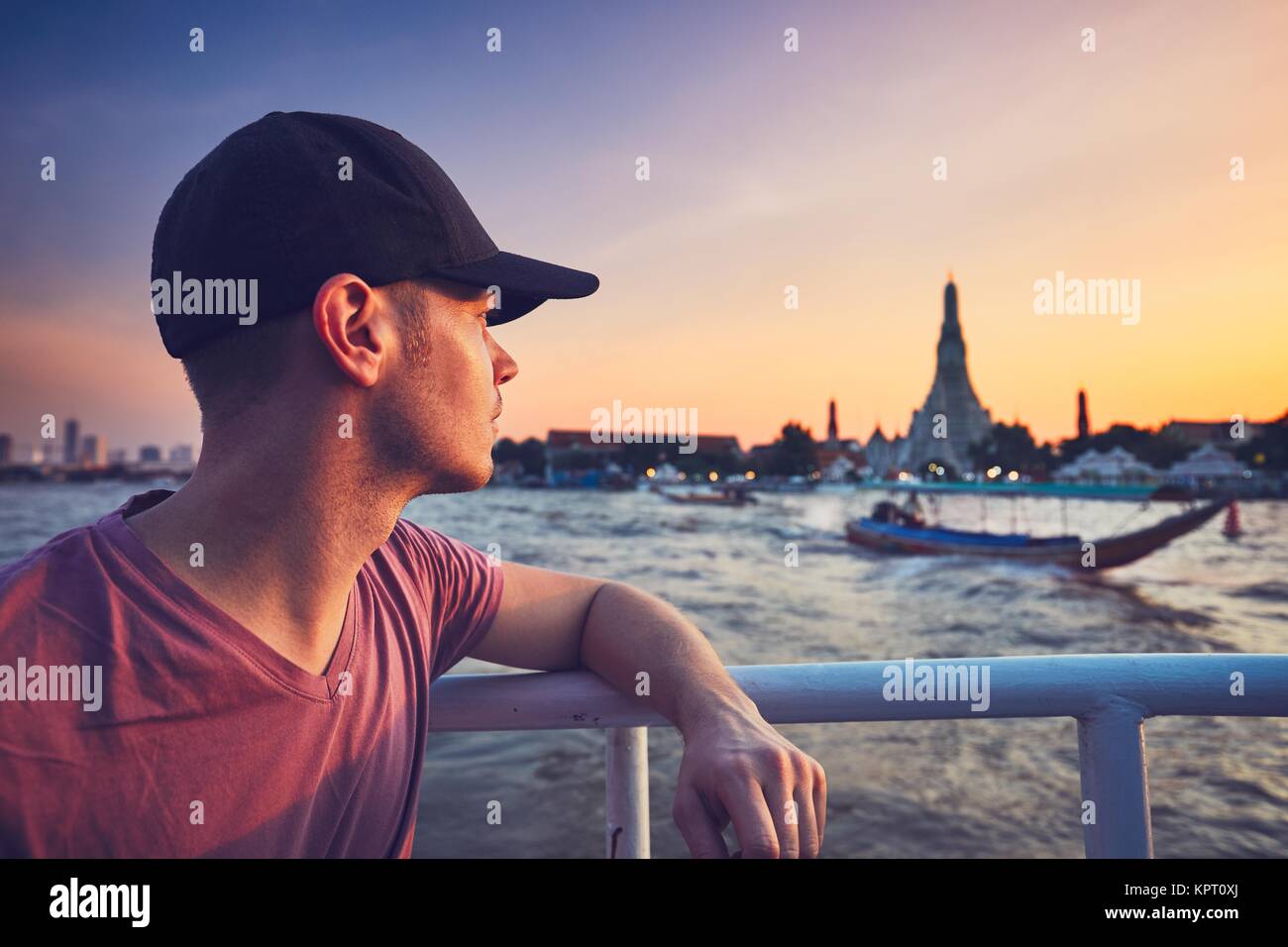 Young man (tourist) on the boat watching the traditional boats against temple Wat Arun. Bangkok at the sunset, Thailand. Stock Photo