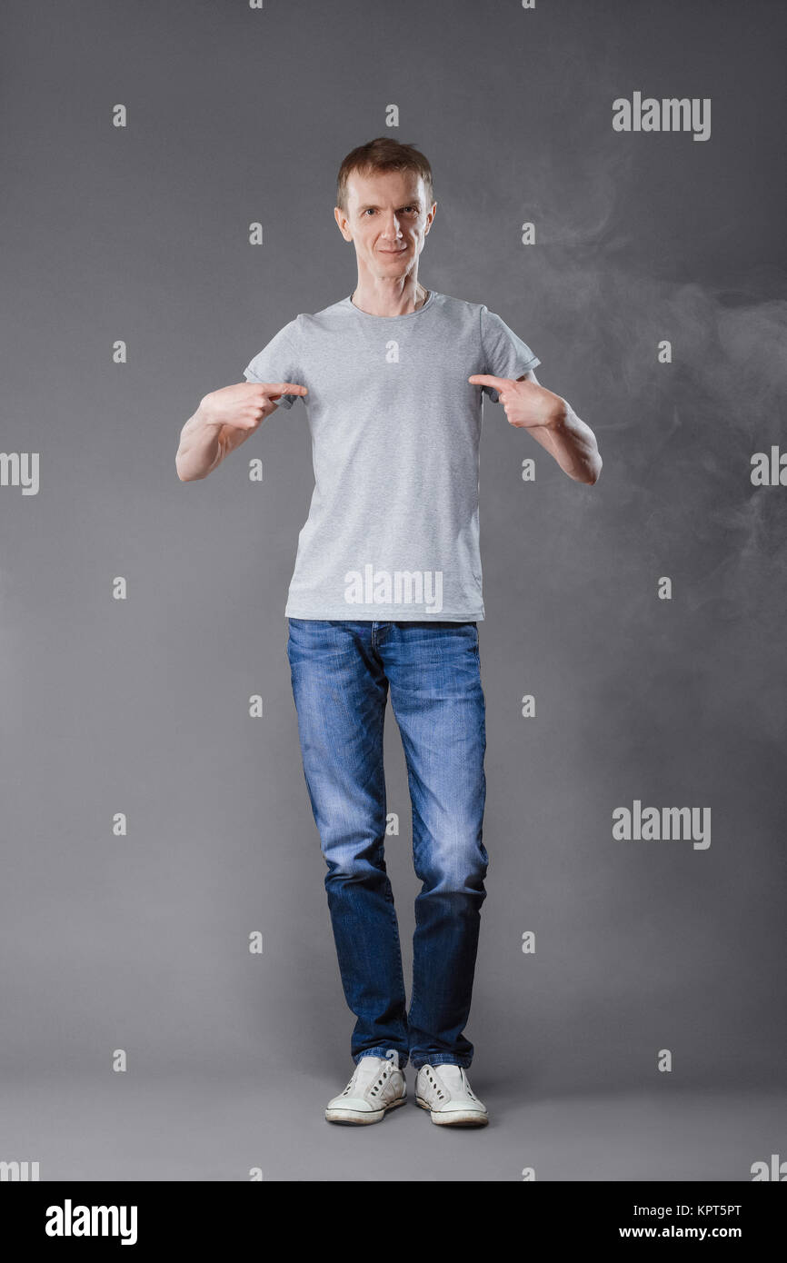Middle age man in jeans pointing fingers to his t-shirt on gray background. Stock Photo