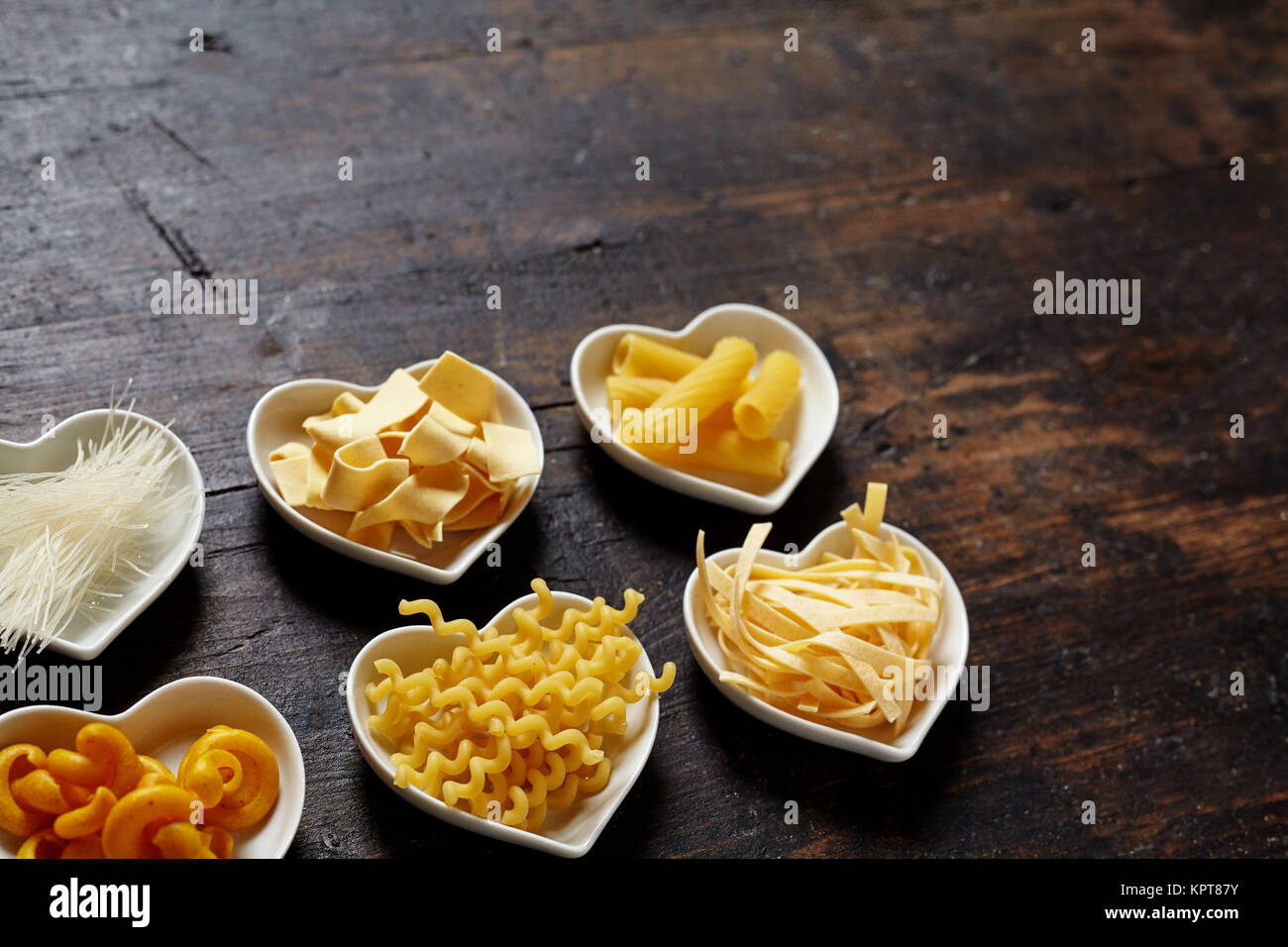 Kleine Herz Schalen gefÃ¼llt mit Nudeln, sortiert. Nudelsorten arrangiert auf dunklem braunem Holz Untergrund mit Gestaltungsfreiraum oder Textfreiraum fÃ¼r italienische Pasta Konzepte. Stock Photo