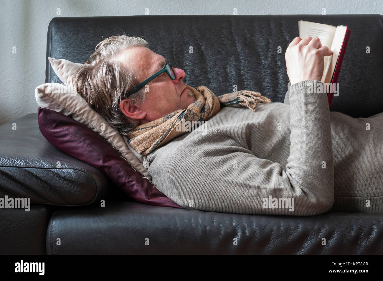 Liegender Mann auf einem Sofa mit Brille und Schal liest in einem Buch. Stock Photo