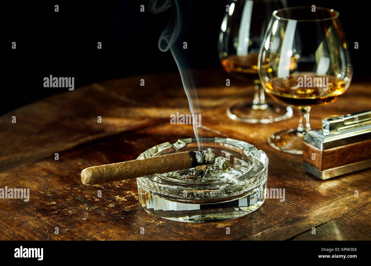 Glass ashtray with smoking cigar by two glasses filled with bourbon