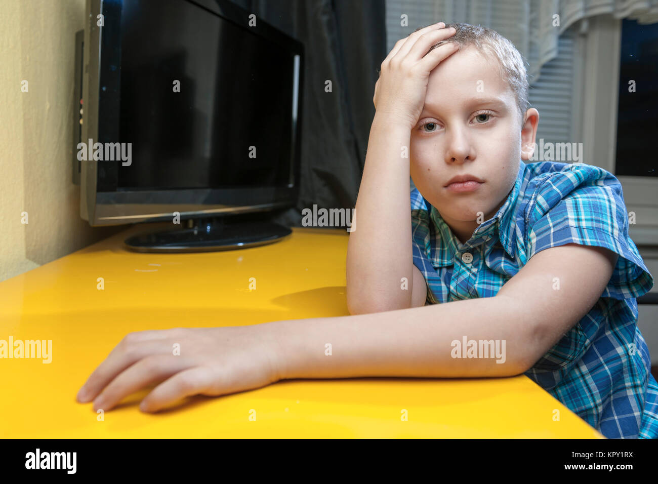 Oberkörper-Ansicht eines zehnjährigen Jungen im blau karierten kurzärmilgen Hemd den linken Arm auf eine gelbe Tischplatte mit einem TV im Hintergrund liegend. Die rechte Hand am Kopf. Ernster Blick in die Kamera. Stock Photo