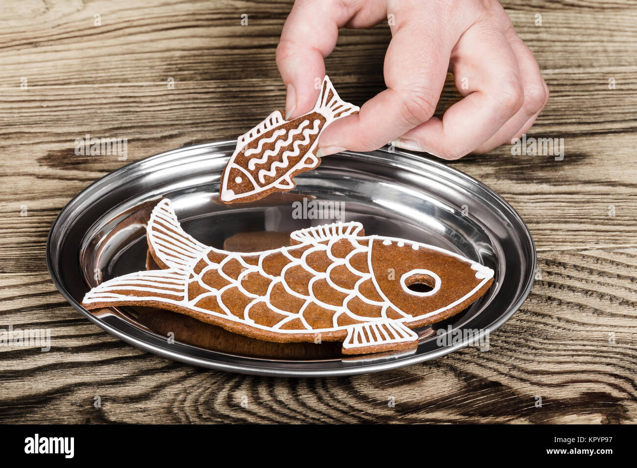 Small and big fish. Christmas gingerbreads on a silver tray. Female hand holding the cute sweet biscuit above the glossy plate on wood background. Stock Photo