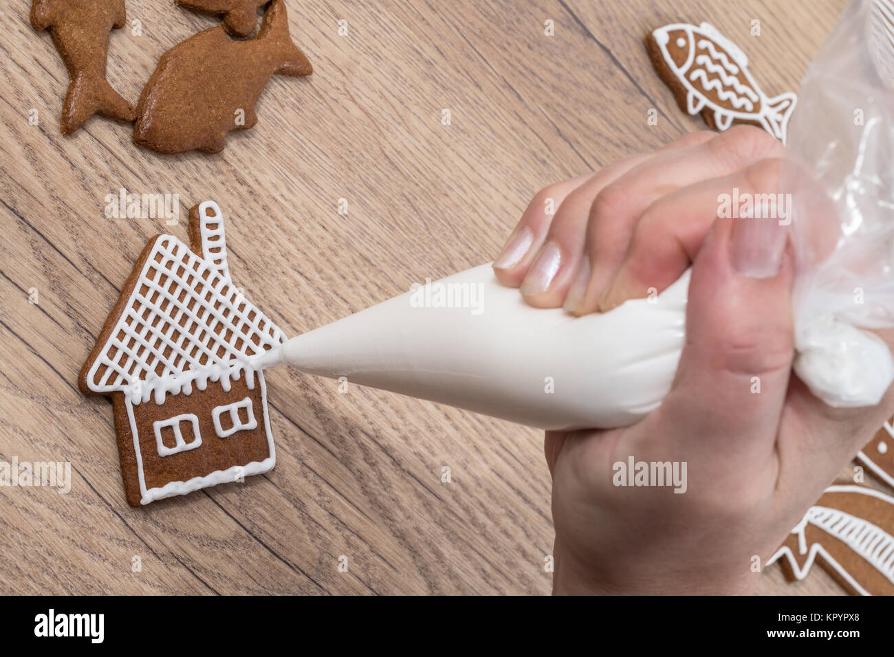 Close-up of female hand while decorating Christmas gingerbread on wood. The woman is painting the house on biscuit using icing bag with sugar frosting Stock Photo