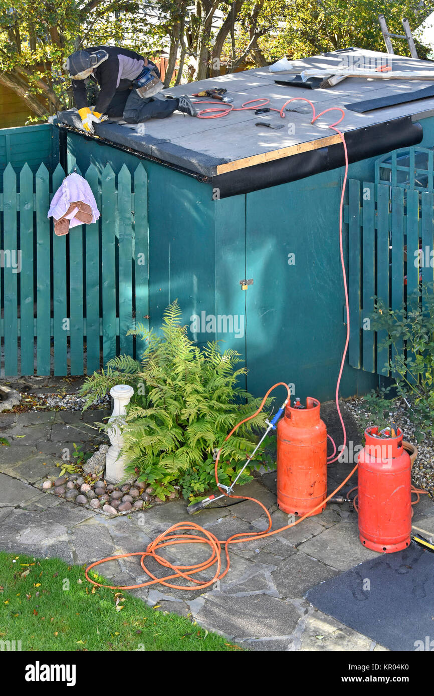 Roofer working on re-covering leaking flat garden shed roof pressing down on heated bitumen adhesive on new roofing felt propane gas cylinders below Stock Photo
