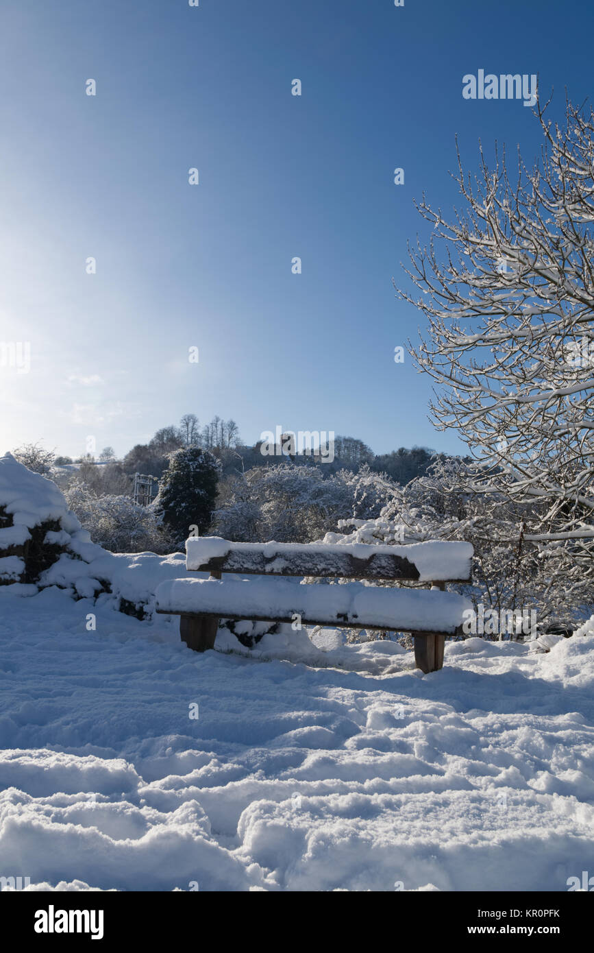 Winter snow covered wooden bench on canal in Kinver, Staffordshire with trees and church in the background Stock Photo