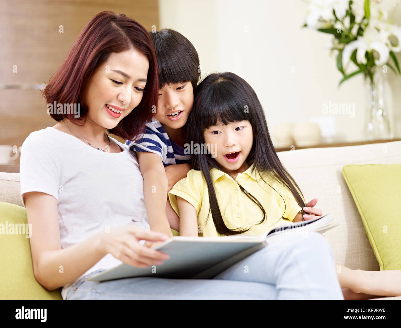 young asian mother sitting on couch at home reading story to two little children. Stock Photo