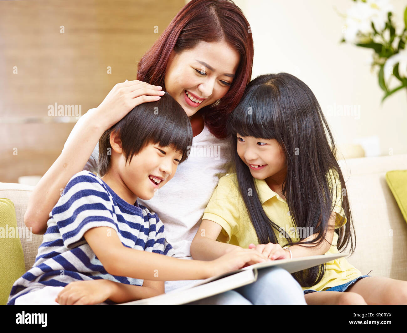 young asian mother sitting on couch at home reading story to two little children. Stock Photo