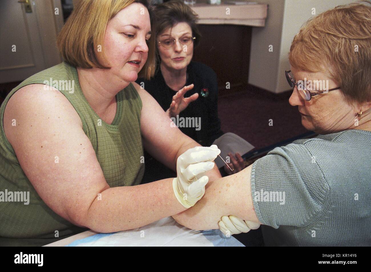Lorna Will (Left) and Jacqueline Kowalski practice proper technique with a bifurcated needle as trainer Judy Gibson observes, 2002. In December 2002, CDC Clinicians trained state licensed vaccine administers how to deliver smallpox vaccine safely and efficiently. Once training was completed, they provided additional smallpox vaccine administration training in their home states. Image courtesy CDC. Stock Photo
