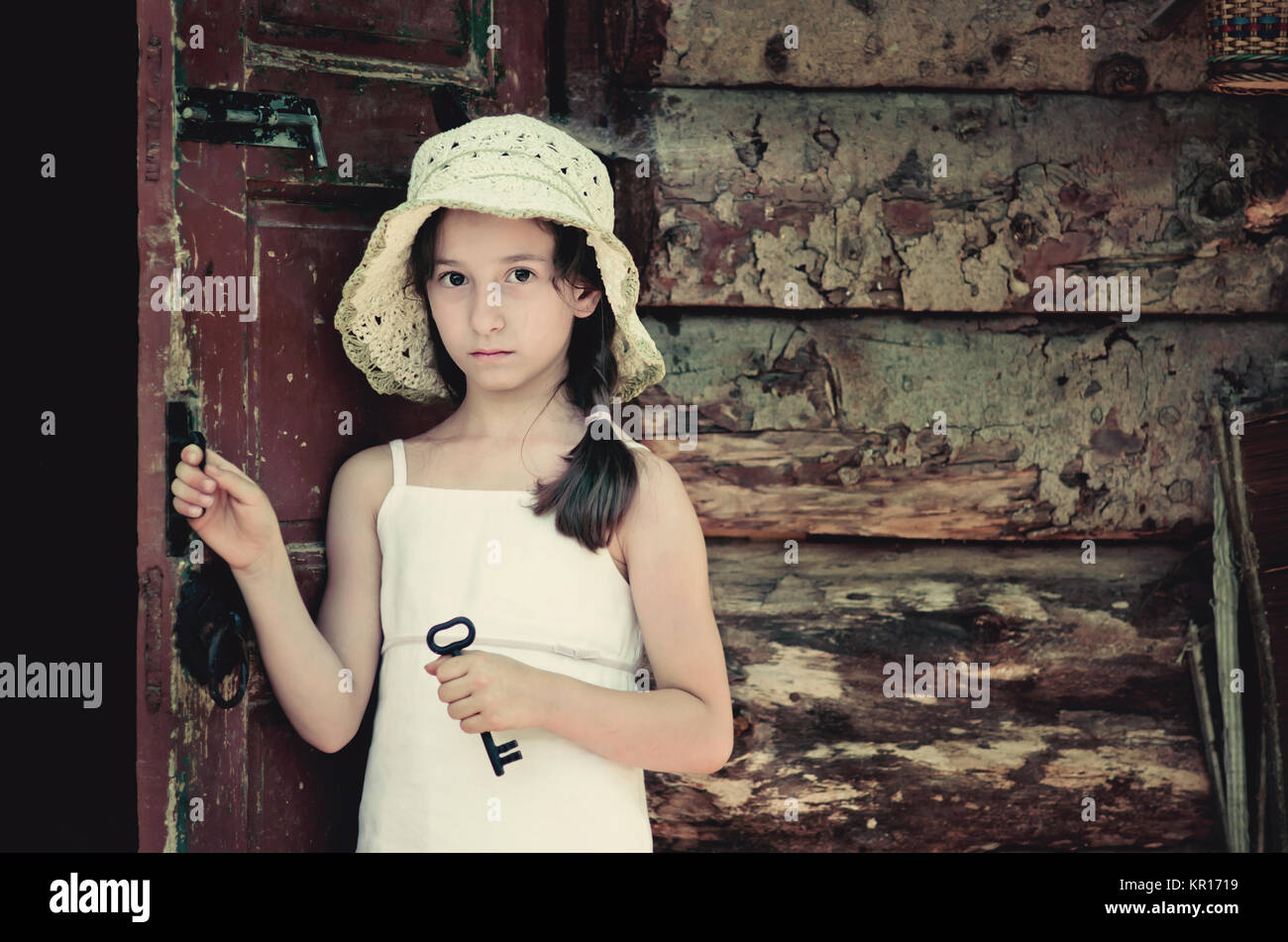 Pretty young girl standing at the door outside an old wooden hut and holding a key. Retro and vintage look, nostalgic mood. Stock Photo