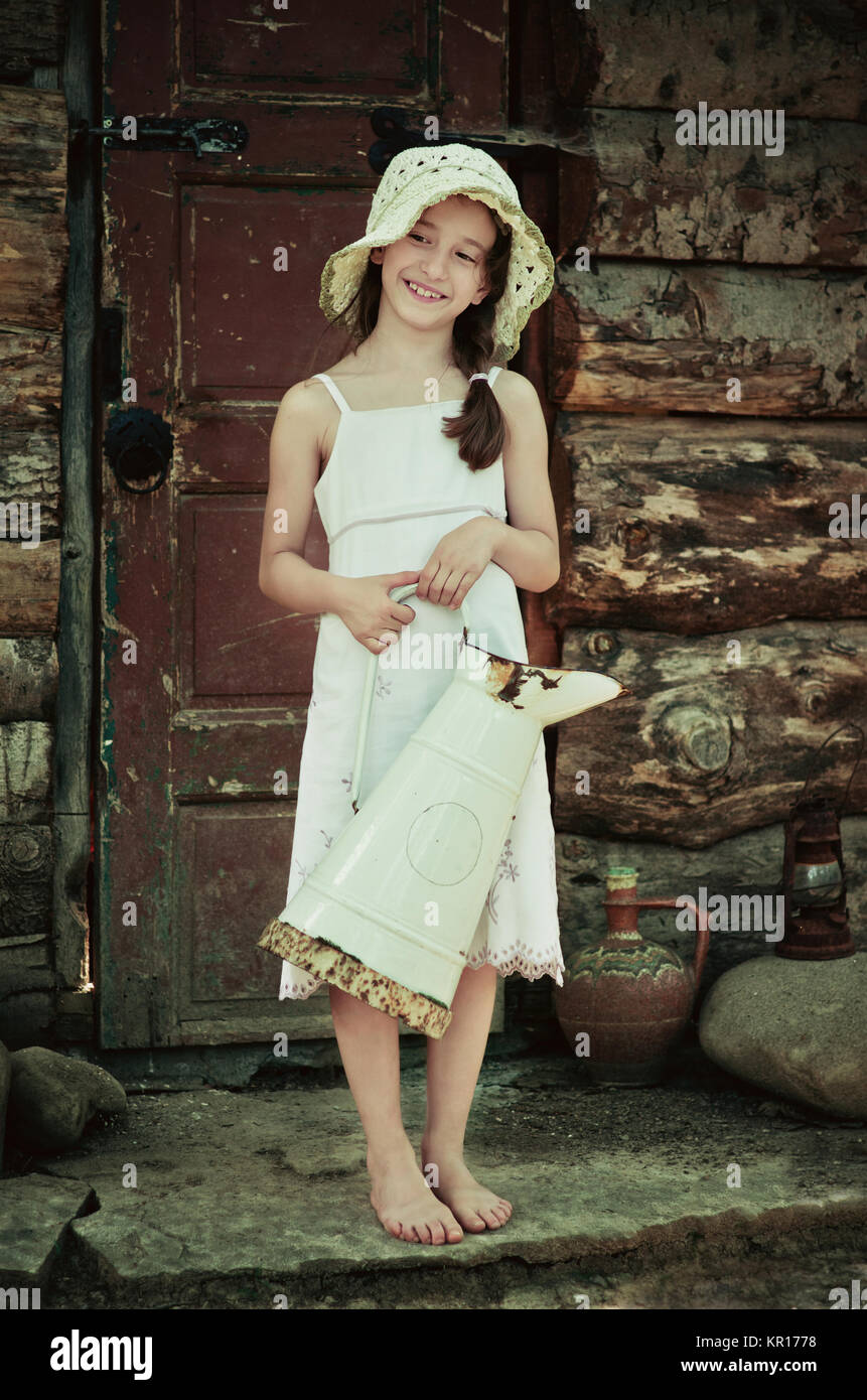 Young girl 5-7 years holding a jug and standing in front of an old wooden house. Retro and vintage looking image. Stock Photo