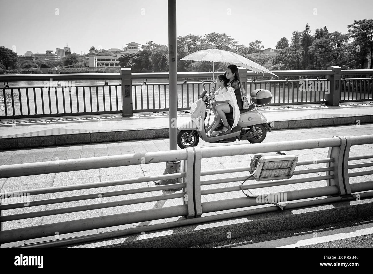 Guilin, China - September 16, 2017: Woman with a child rides a scooter and holds umbrella protecting from the sun. Stock Photo