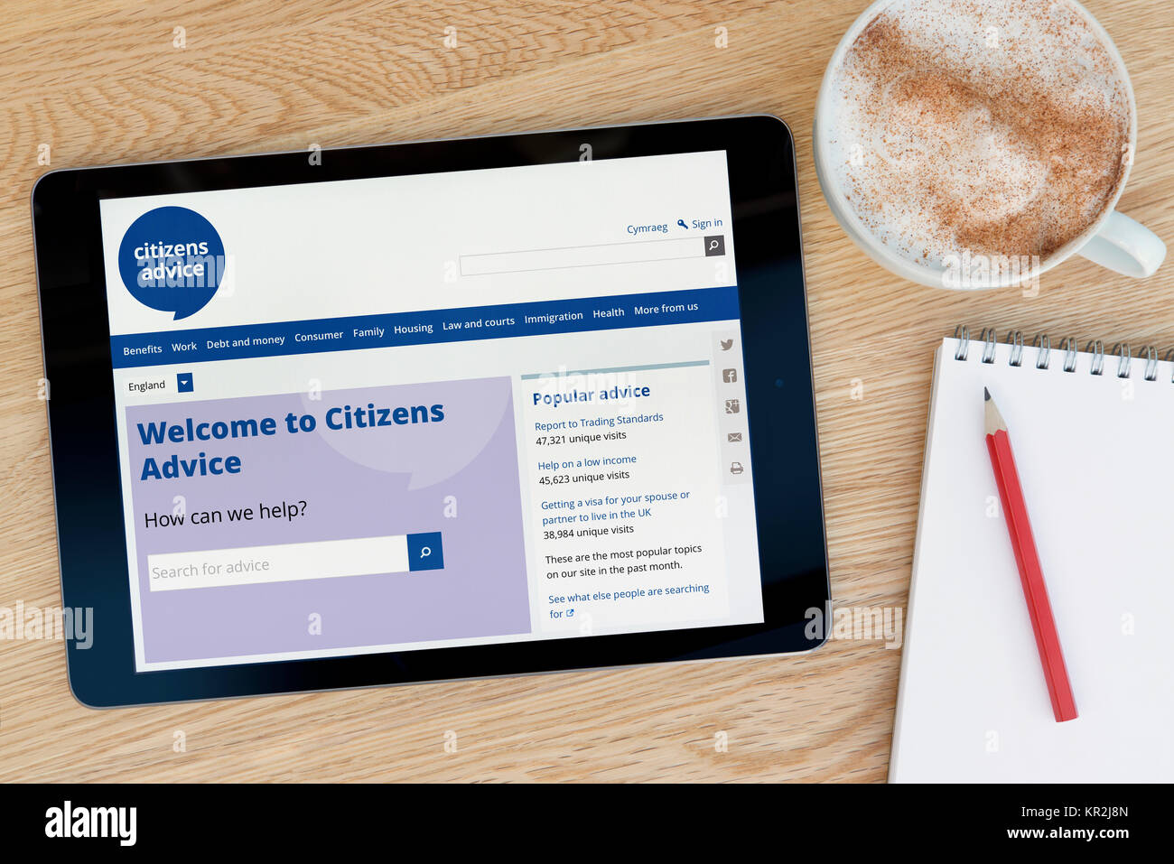 The Citizens Advice website on an iPad tablet device which rests on a wooden table beside a notepad and pencil and a cup of coffee (Editorial only) Stock Photo
