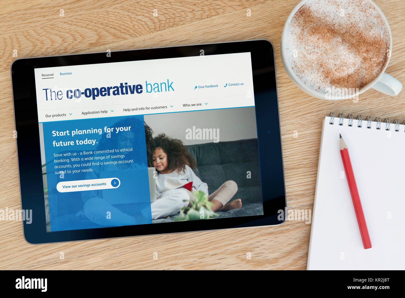 The Co-operative Bank website on an iPad tablet device which rests on a wooden table beside a notepad and pencil and a cup of coffee (Editorial only) Stock Photo