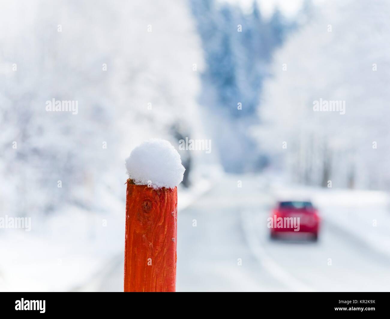 Wooden post with patch of snow on top and passing by departing Red car in background blurry snowy Winter road in country-side Stock Photo