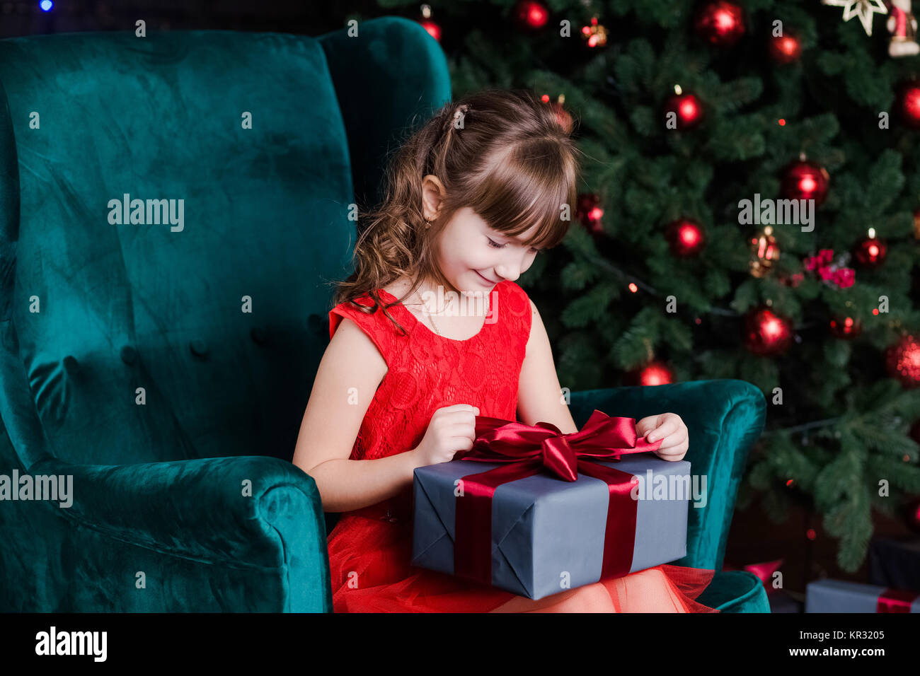 Cute little happy girl with present box sitting in chair near Christmas ...