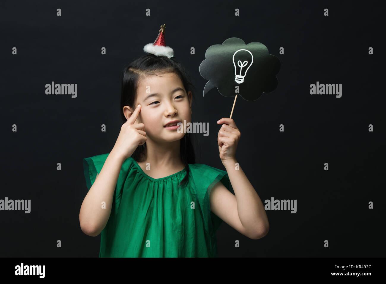 cute asian girl child dressed in a green dress holding an idea bulb stick with a christmas head decoration on a black background. A thinking expressio Stock Photo