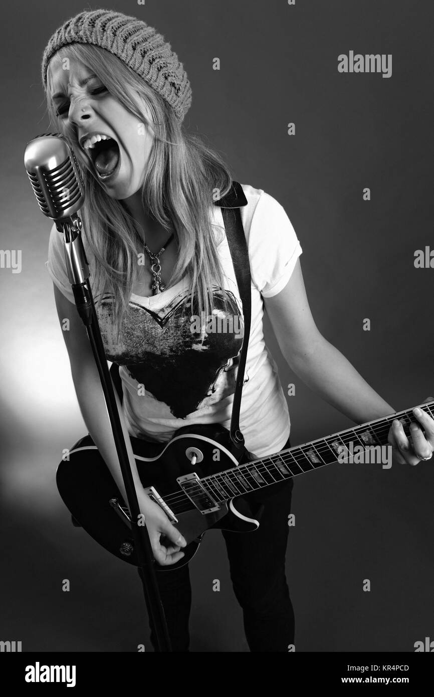 Black and white photo of a blond female screaming into an old microphone and playing electric guitar.  High contrast with film grain added. Stock Photo