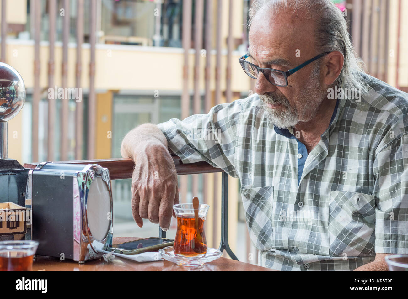 Old Turkish man with eyeglasses looking at his smartpone on the table in a restaurant. Traditional red Turkish tea is ready after the meal. Stock Photo