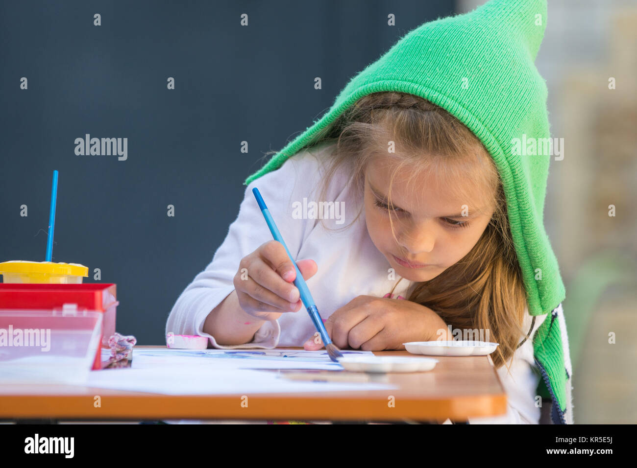 Girl enthusiastically paints watercolors sitting at a table in the yard ...