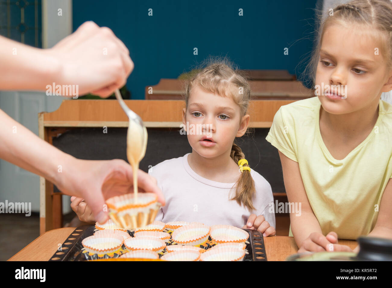 Two girls with a surprised look like mom pours batter into molds for cupcakes Stock Photo