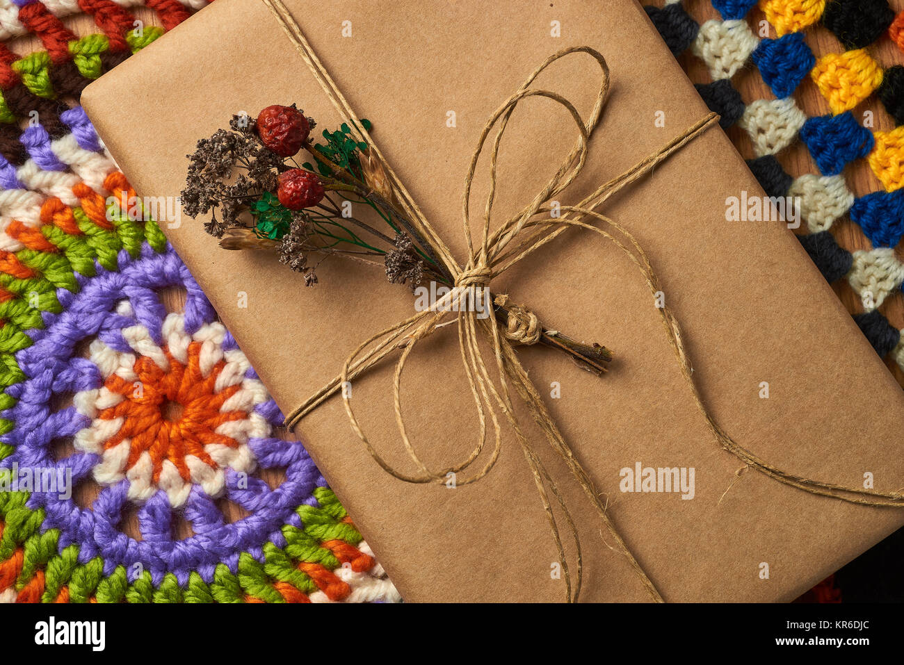Wrapped book tied with brown paper thread with small dried bouquet of field plants is lying on the table on two colorful knitted napkins. Stock Photo