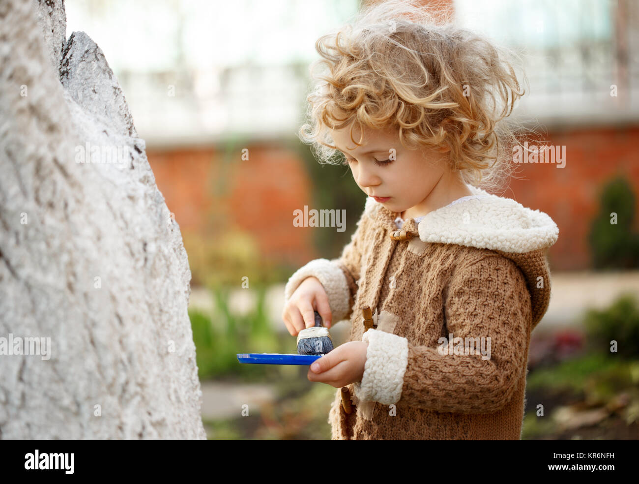Little adorable blonde girl covering the tree with white paint to protect against rodents in the garden in the spring. Stock Photo