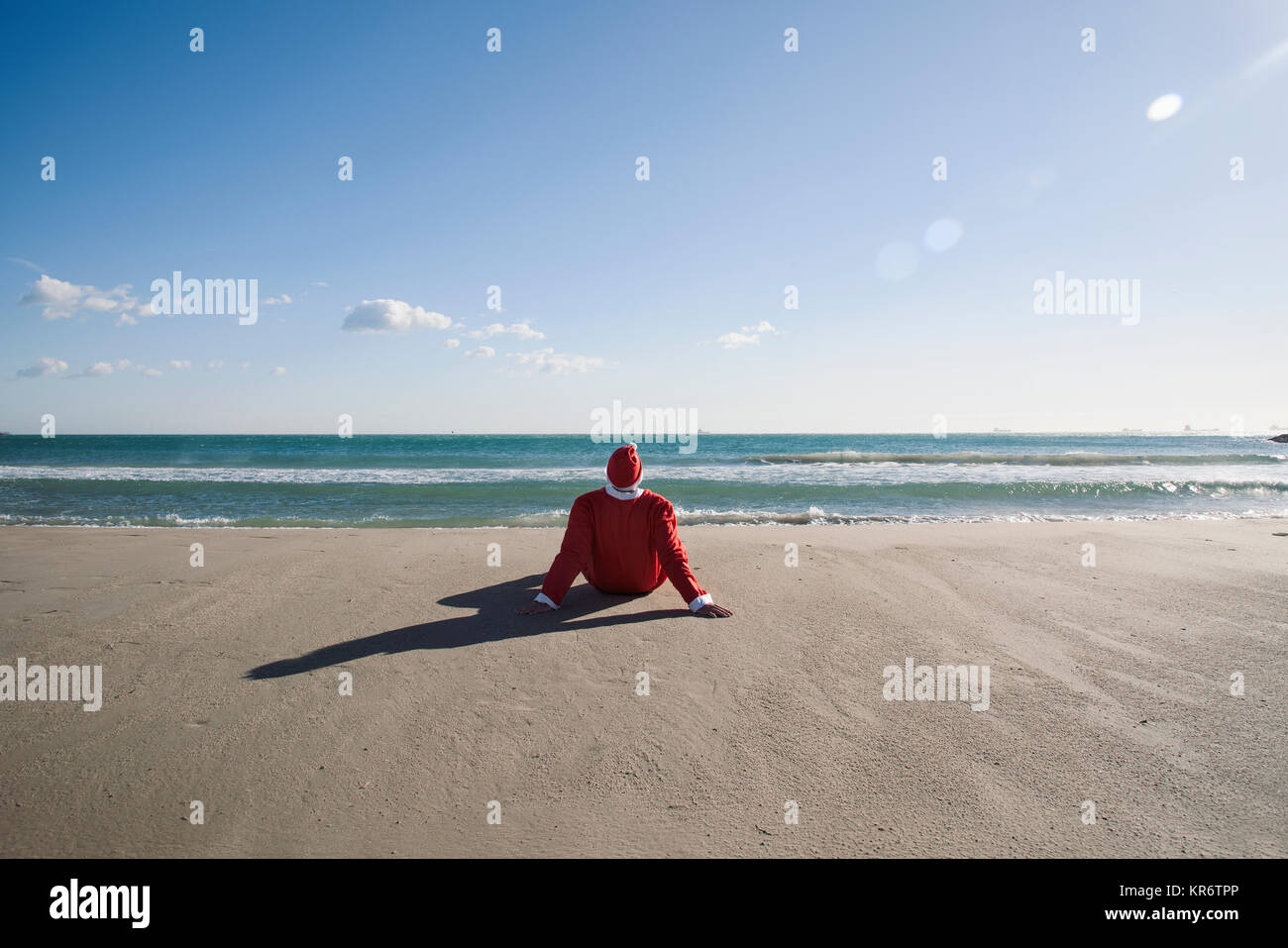 santa claus, seen from behind, sitting on the sand of a beach facing ...