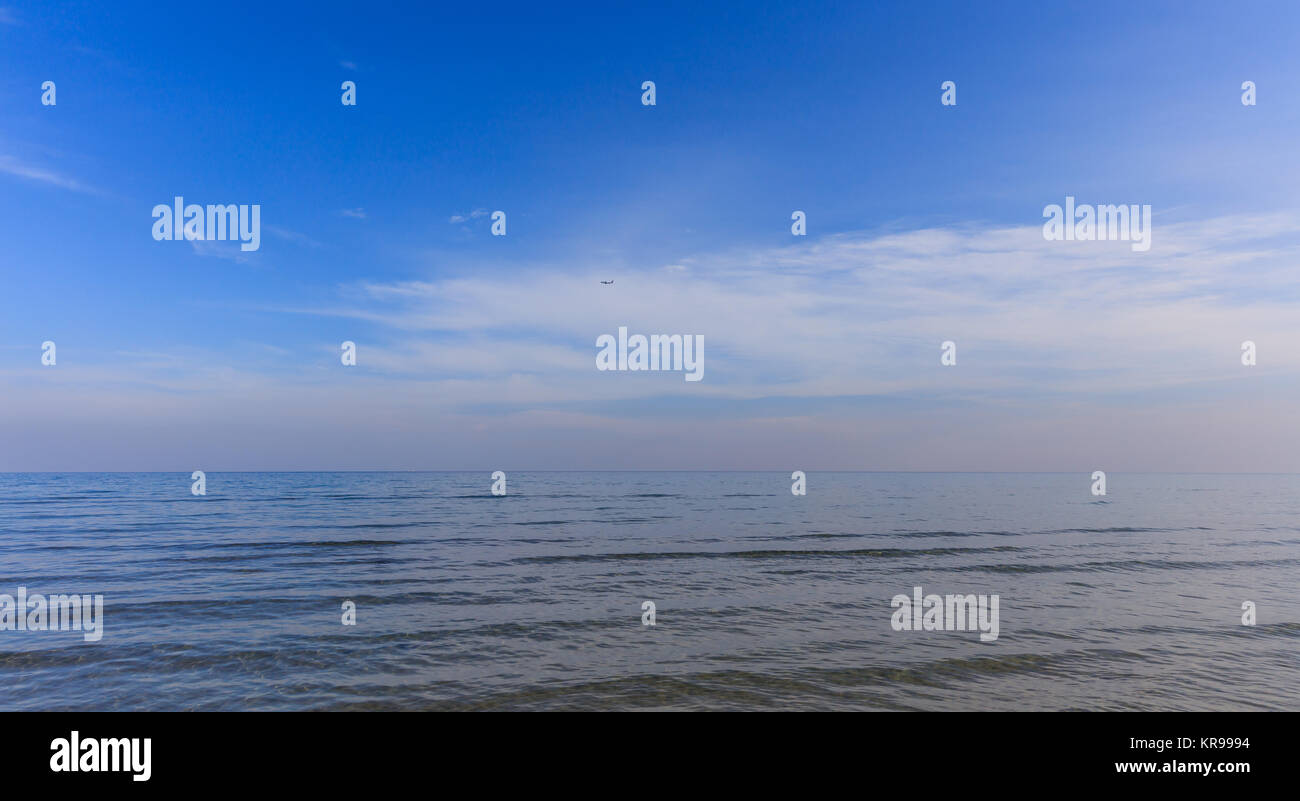 Calm clear sea and blue sky with white clouds background. An airplane fly on the horizon. Stock Photo
