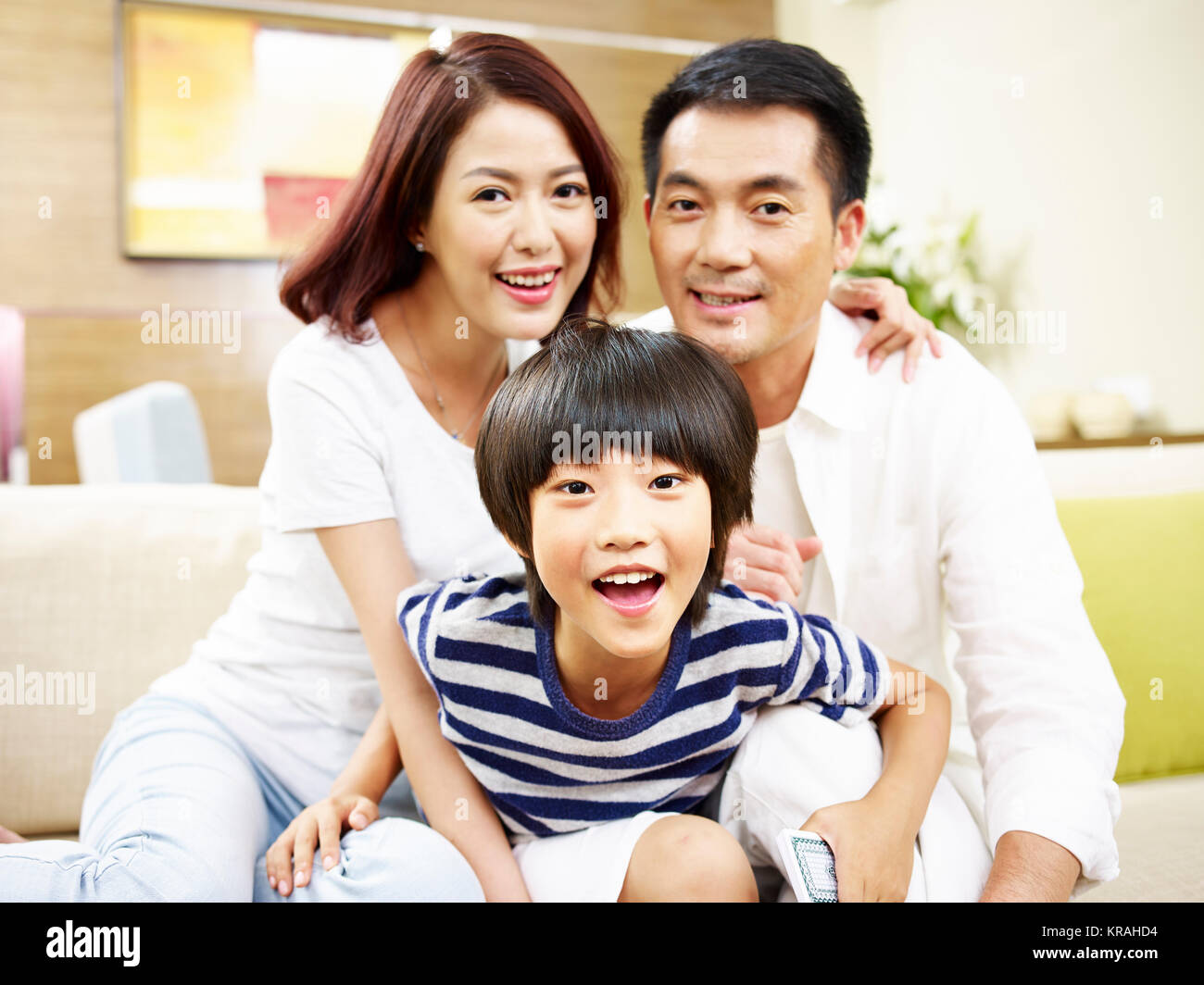 portrait of a happy asian family mother father and son sitting on couch at home looking at camera smiling. Stock Photo