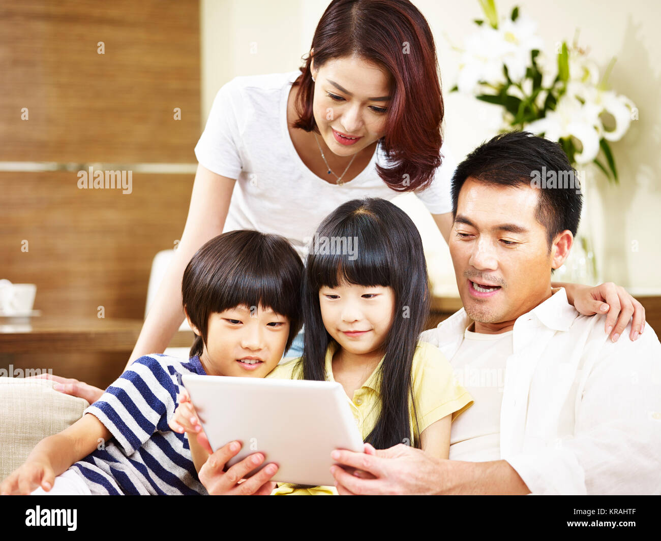 asian parents mother and father and two children son and daughter sitting on family couch using digital tablet together. Stock Photo
