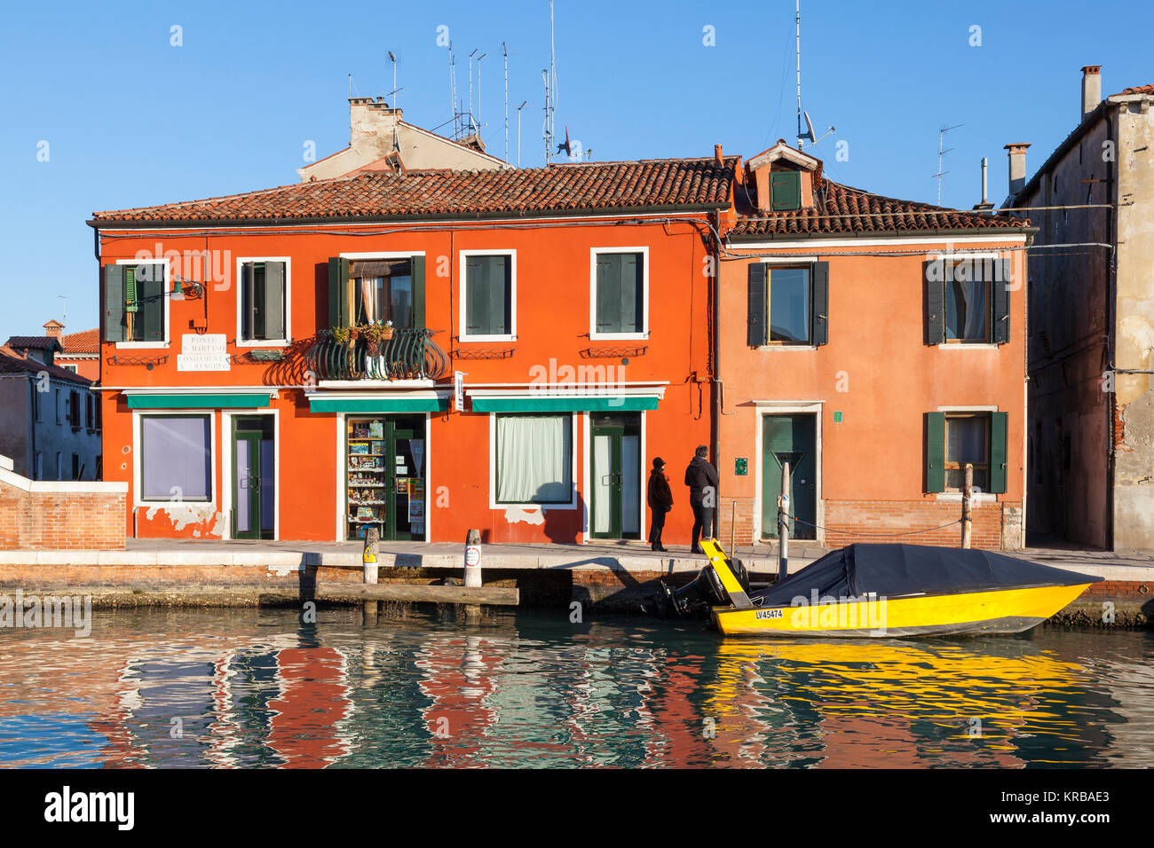 Colorful houses and boat at sunset, Murano Island, Venice, Italy with a couple strolling on Fondamenta A Maschio and reflections in the canal Stock Photo