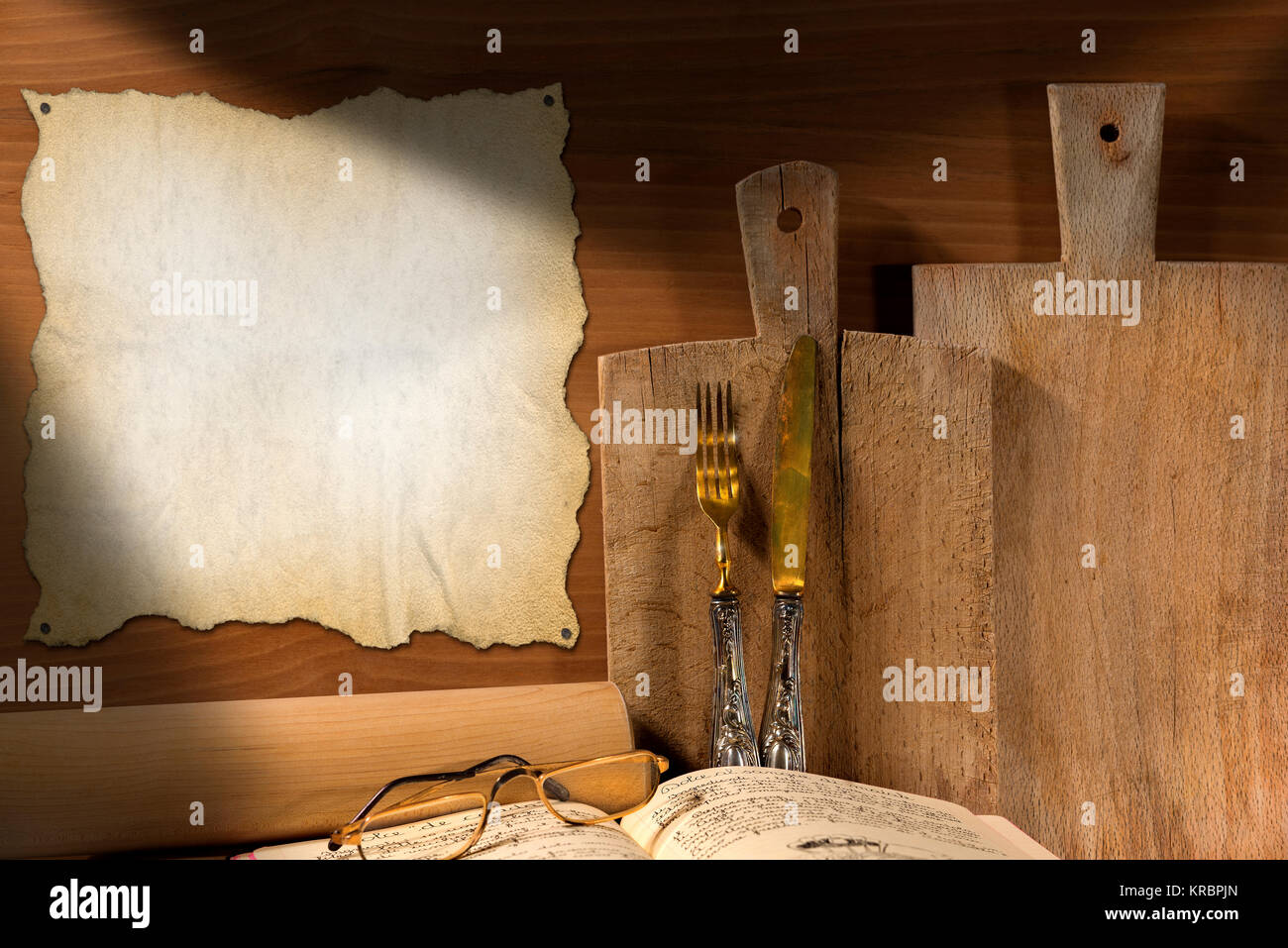 Wooden wall with cutting boards, silverware, recipe book, glasses, rolling pin and empty parchment. Concept of rustic cuisine Stock Photo