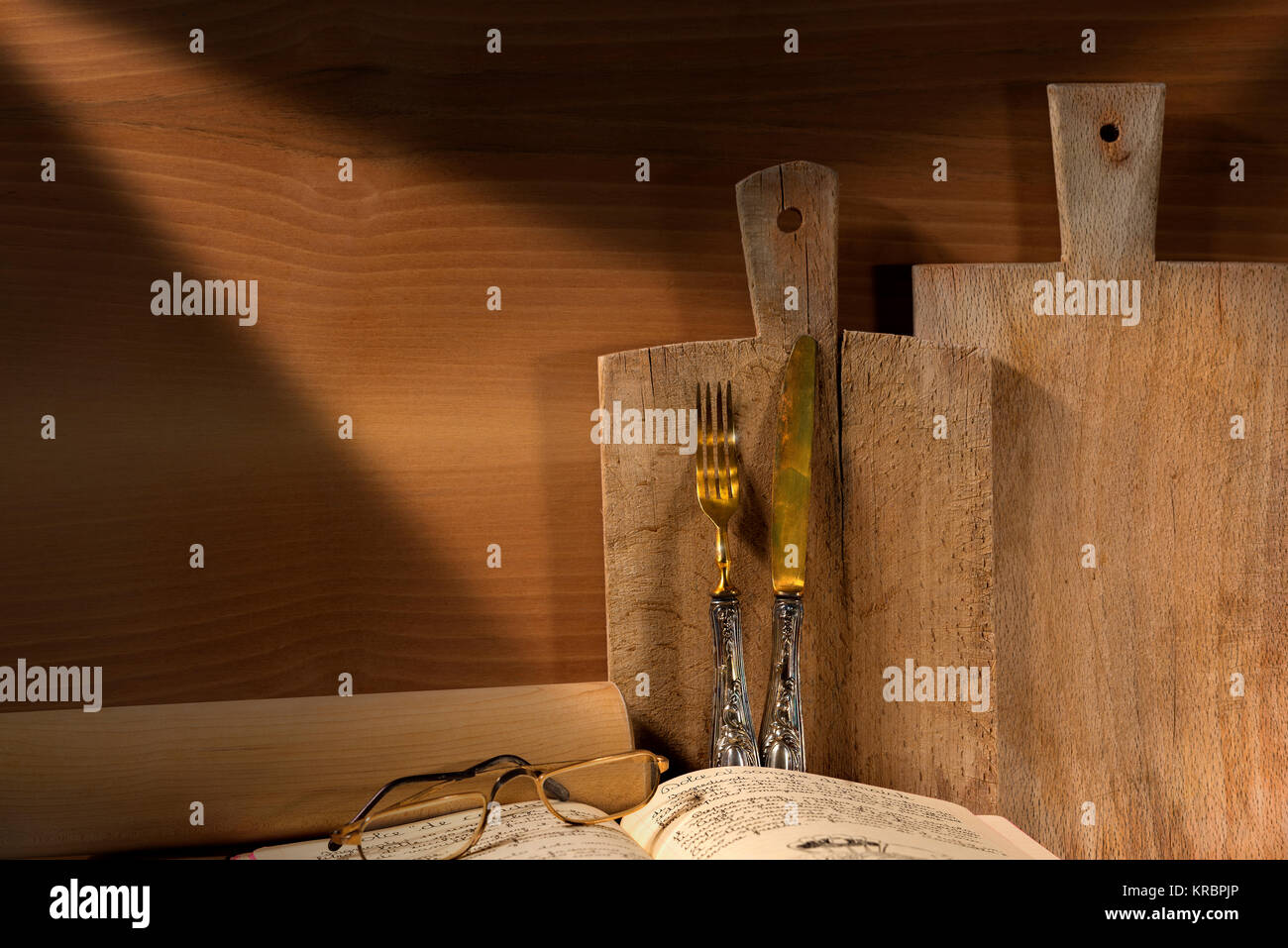 Wooden wall with two cutting boards, old silverware, recipe book, glasses and rolling pin. Concept of rustic cuisine Stock Photo