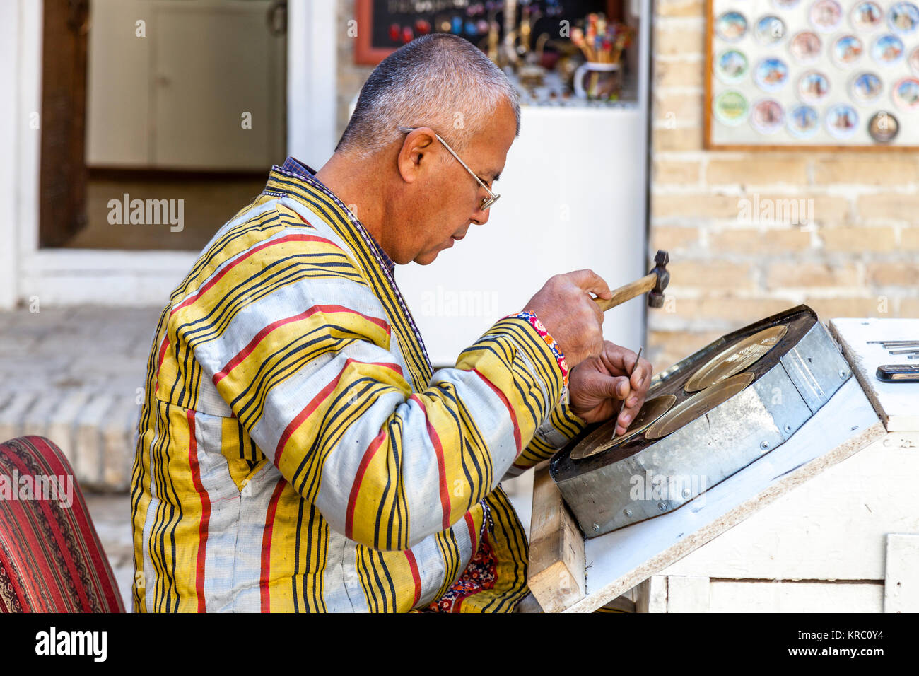 A Local Craftsman In Traditional Dress Engraving A Brass Plate In The Market, Bukhara, Uzbekistan Stock Photo
