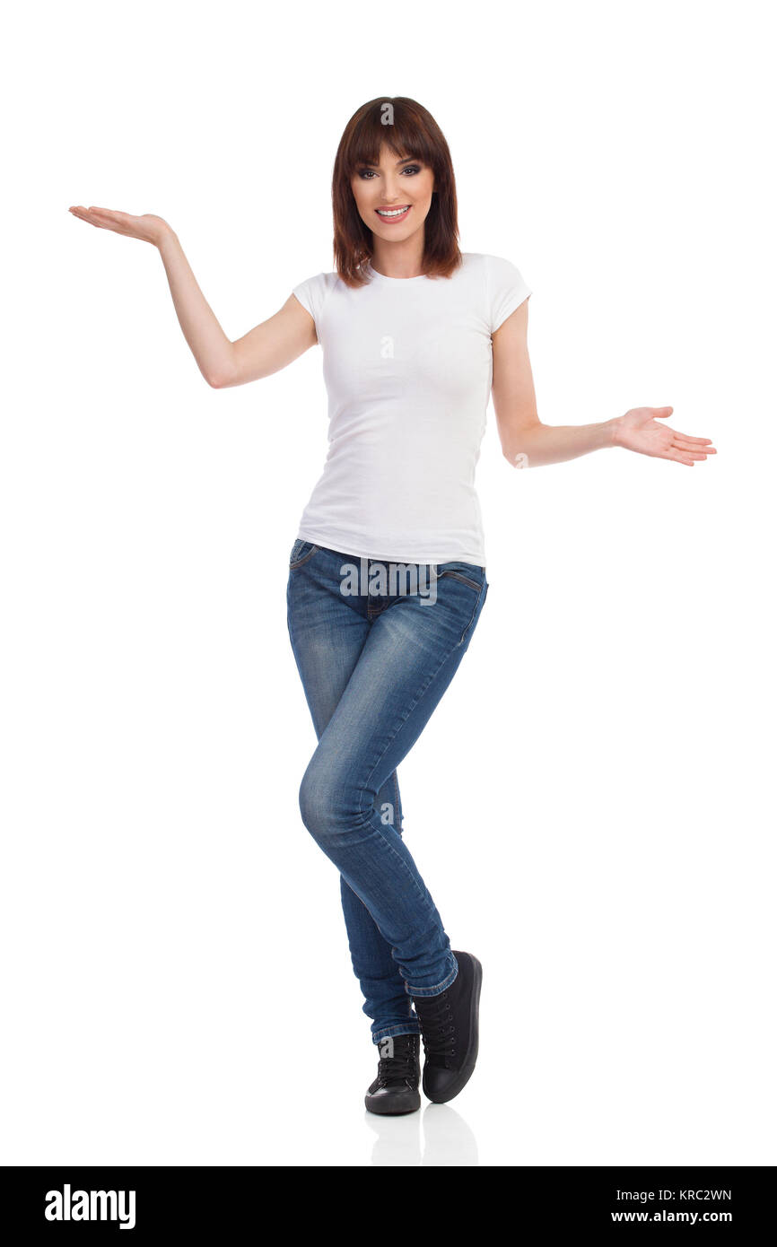 Young woman in jeans, white t-shirt and sneakers is standing with hands raised, looking at camera, smiling and presenting. Full length studio shot iso Stock Photo