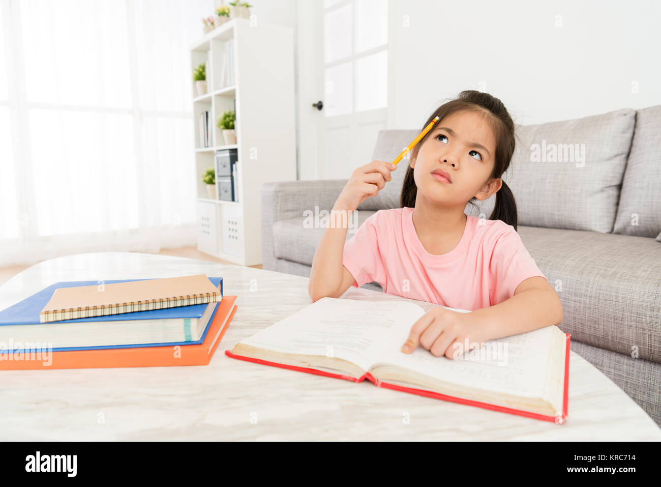 lovely sweet little girl studying school class textbook at home and getting difficulty problem sitting in living room thinking how to solve. Stock Photo