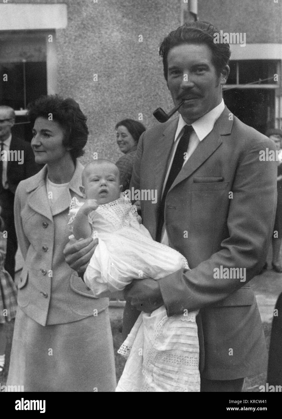 The versatile godfather at a christening manages to hold the baby and smoke his pipe at the same time! Date: circa 1970 Stock Photo