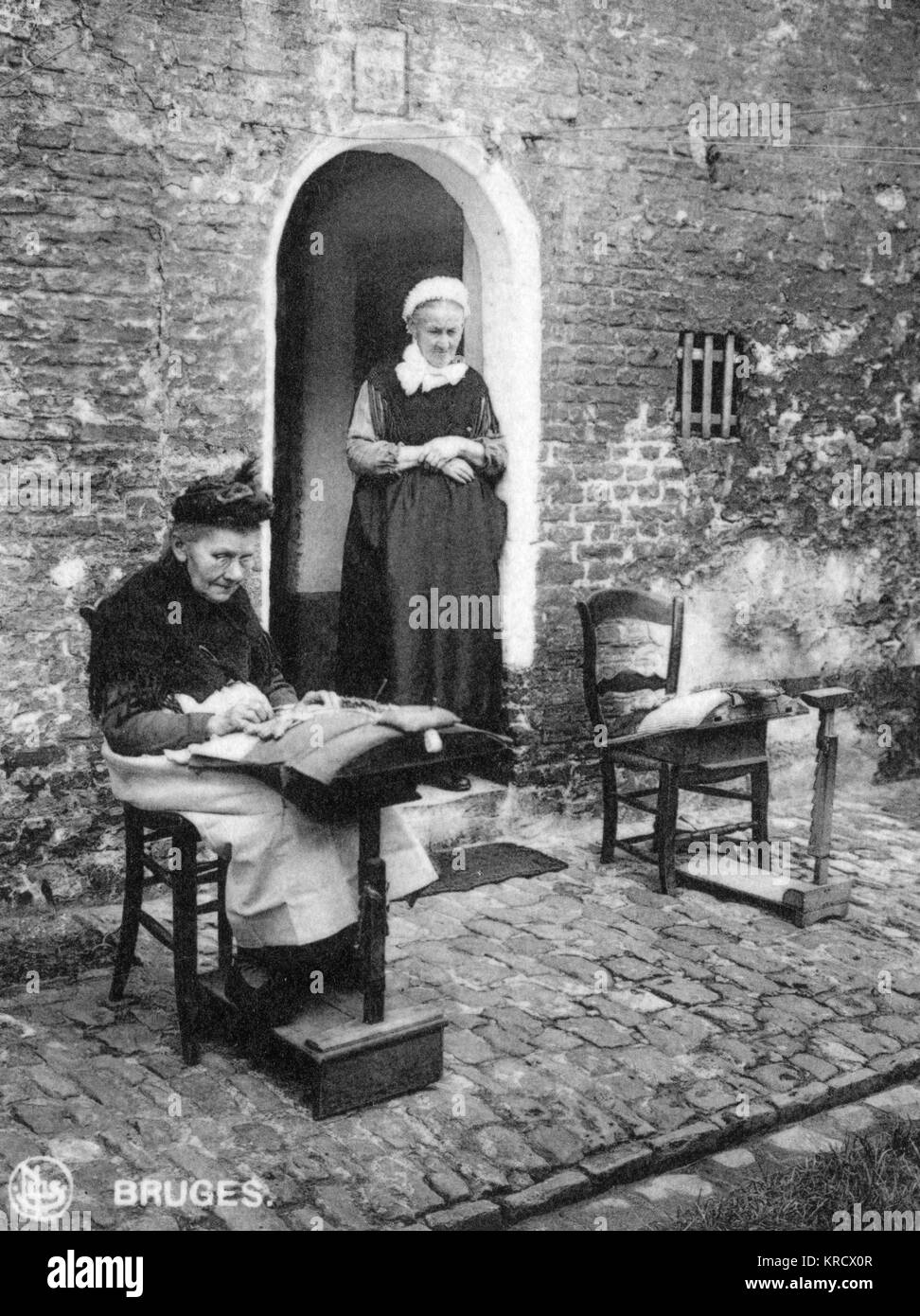 Two elderly women making lace on the pavement outside their home in Bruges (Brugge), Belgium. Date: circa 1920 Stock Photo