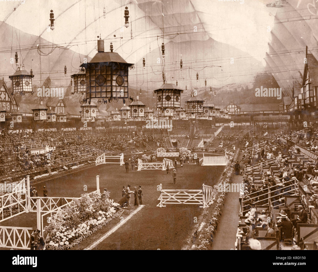 View of the arena inside the Grand Hall at the International Horse Show, Olympia exhibition centre, West Kensington, London.  The arena is set out with fences for the horse jumping events, the auditorium is partly filled with audience members, and large Chinese style lanterns hang from the roof.       Date: circa 1909 Stock Photo