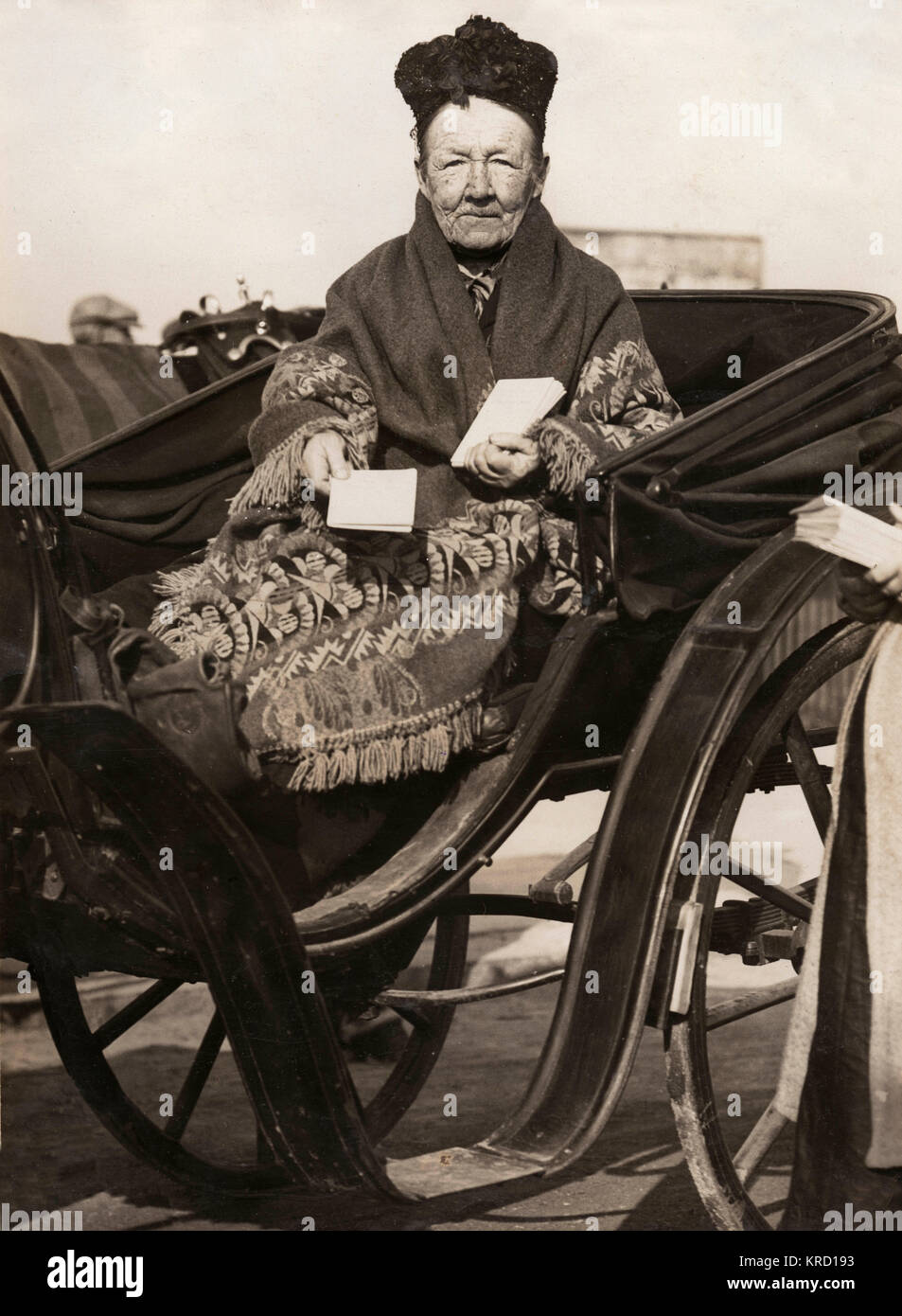 An elderly woman known as Old Kate, who makes her living from the sale of race cards.  She is seen here sitting in an open carriage, provided for her by some of her patrons so that she can continue working despite having recently broken her leg.       Date: circa 1910 Stock Photo
