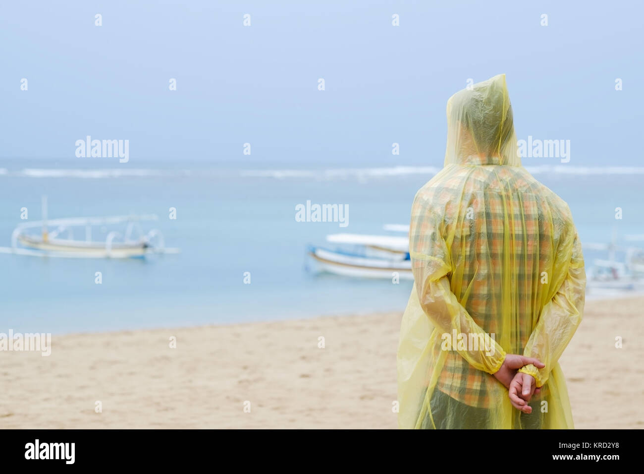 Man in a yellow raincoat on tropical seashore during rain. He is thinking about something in loneliness Stock Photo