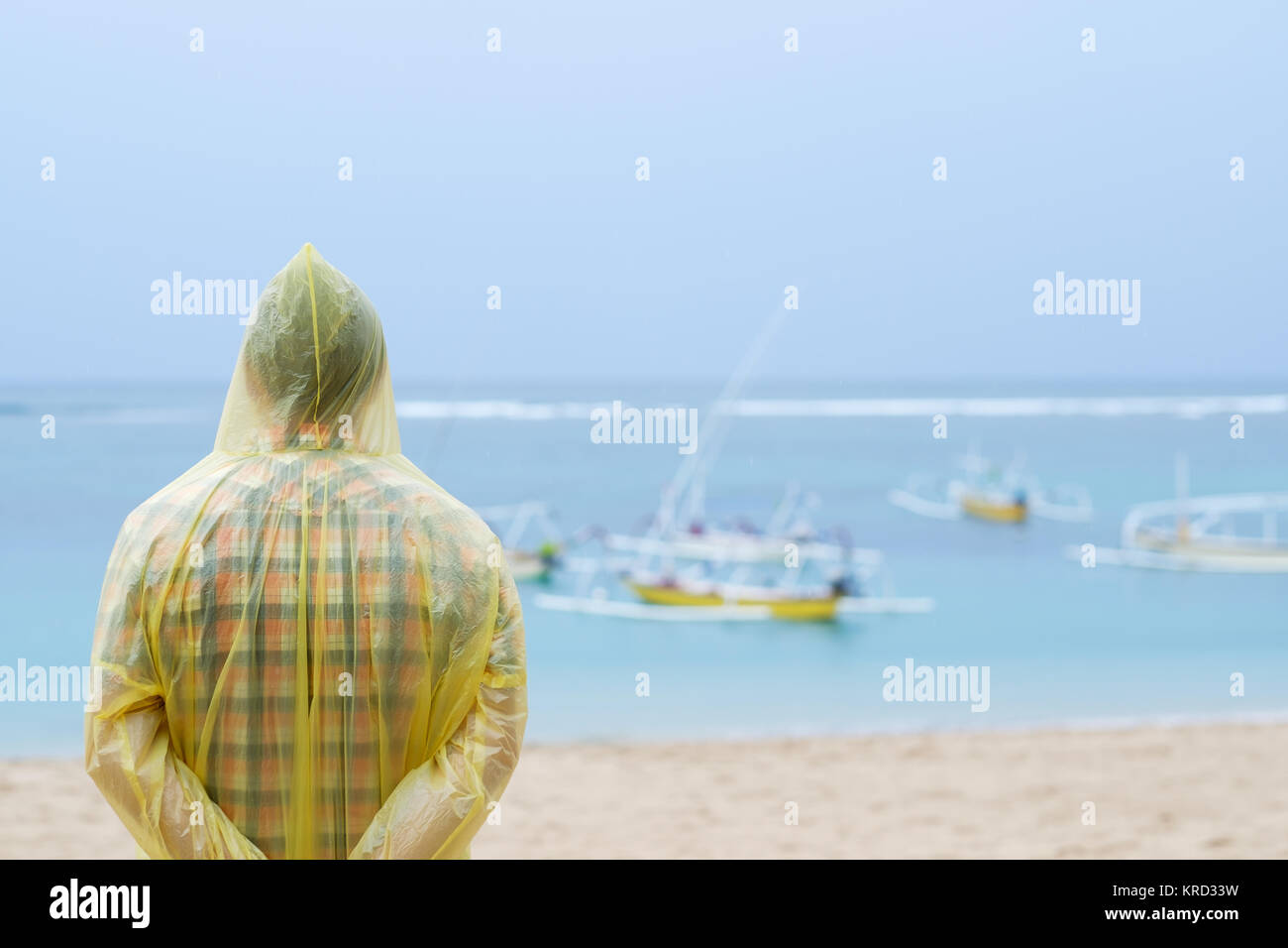 Man in a yellow raincoat on tropical seashore during rain. He is thinking about something in loneliness Stock Photo