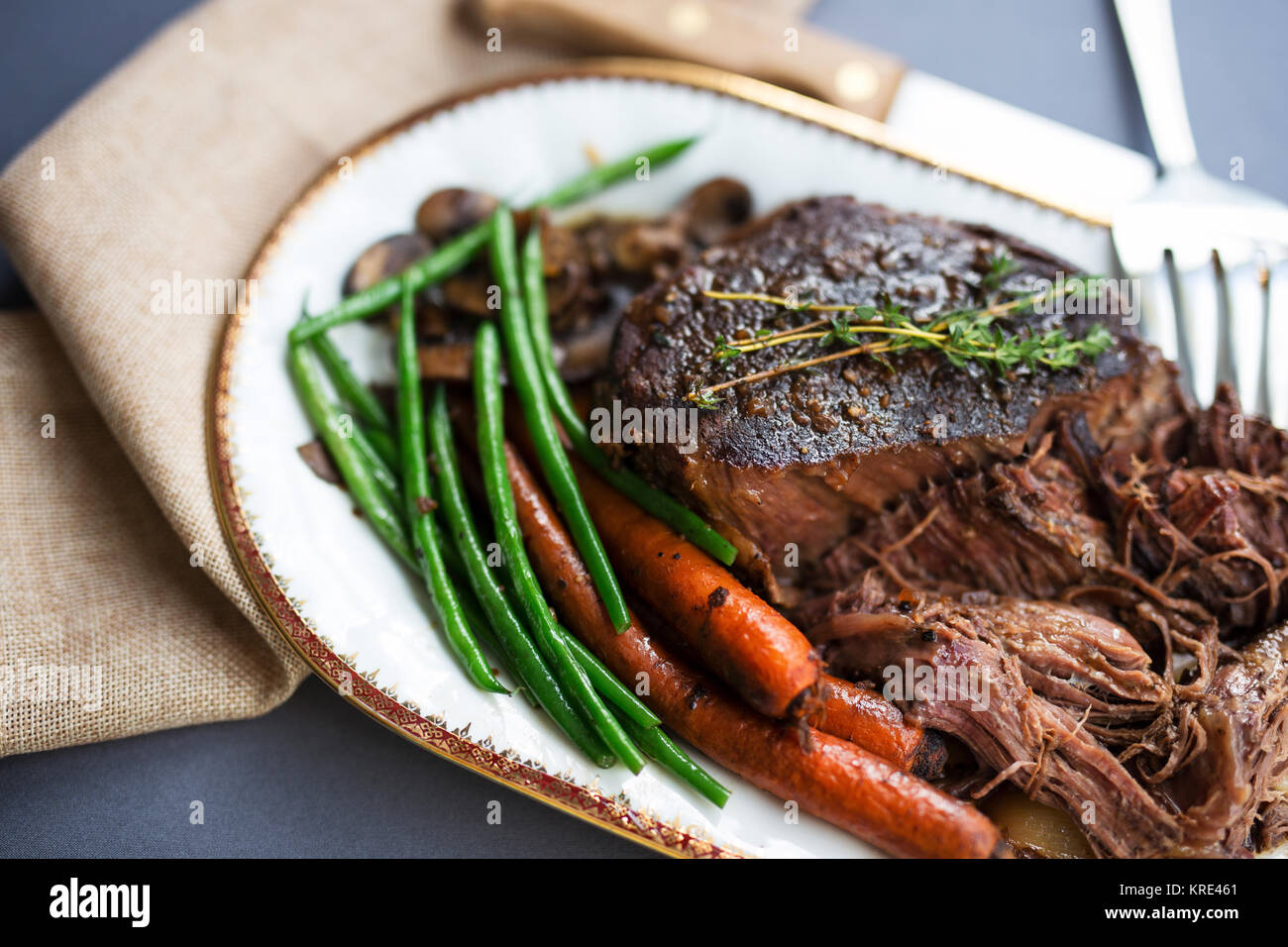 Close up shot of a pot roast with carrots, green beans and mushrooms on a white platter with gold rim. Stock Photo