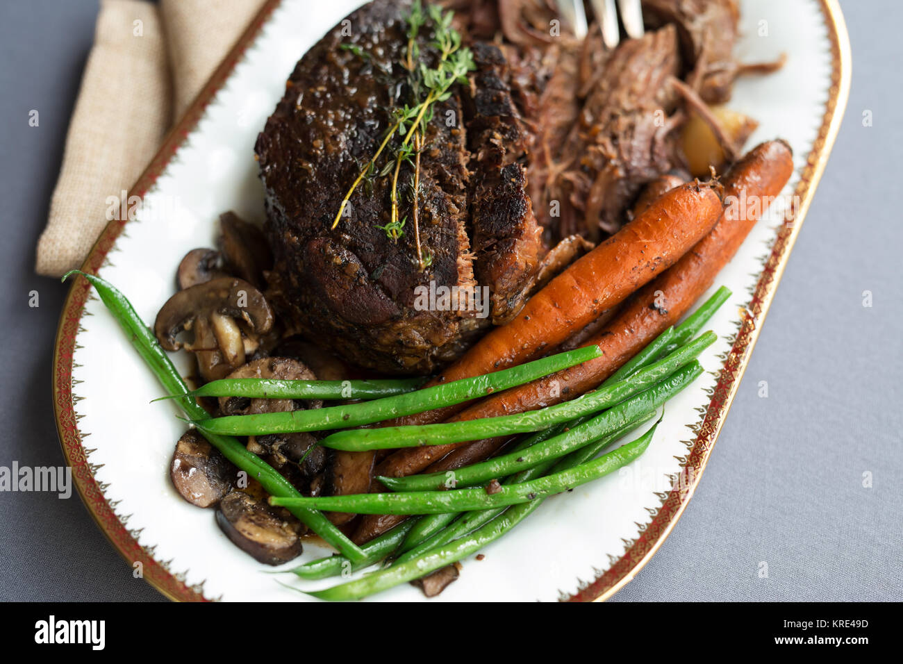 Close up shot of a pot roast with carrots, green beans and mushrooms on a white platter with gold rim. Stock Photo