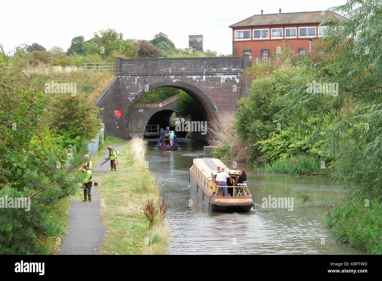 Approaching railway junction bridges, Droitwich Barge Canal, Droitwich, Worcestershire.  With barges on the water, and men cutting back the undergrowth on the left-hand path. Stock Photo