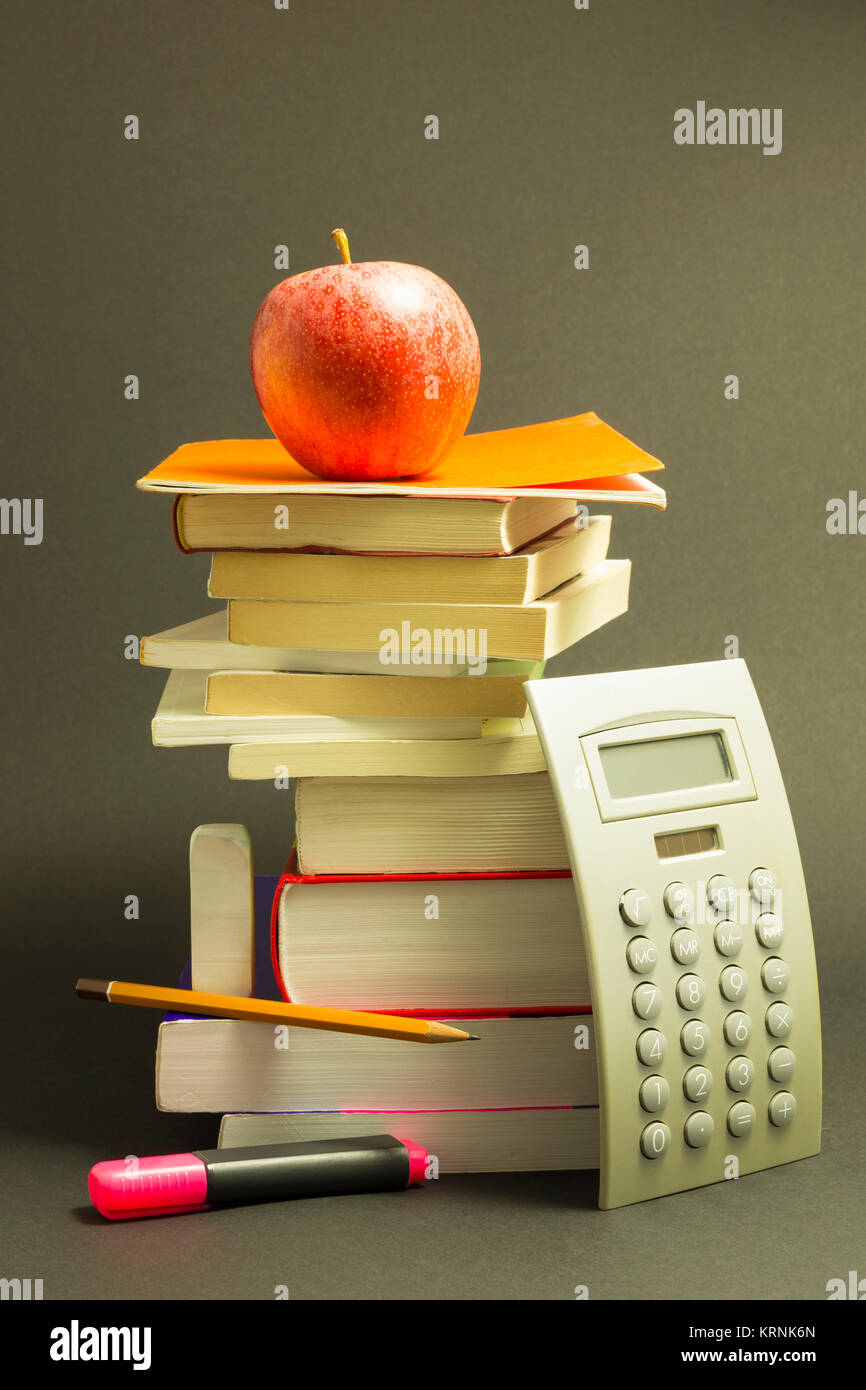 Concept of education. Stack of school books with calculator, pink marker and pencil and a red apple on top in front of dark gray background Stock Photo