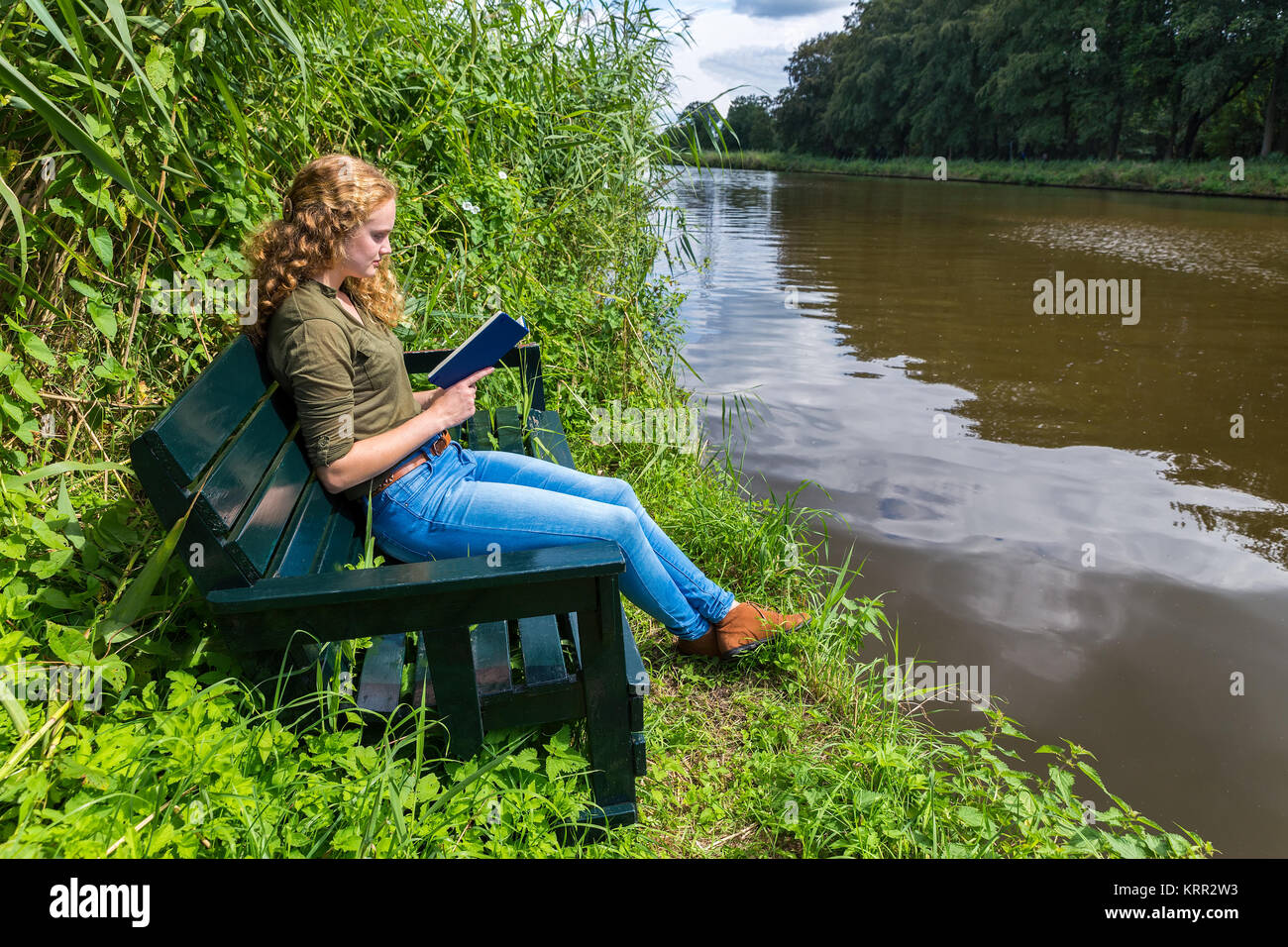 Young caucasian woman reading book on wooden bench at river Stock Photo