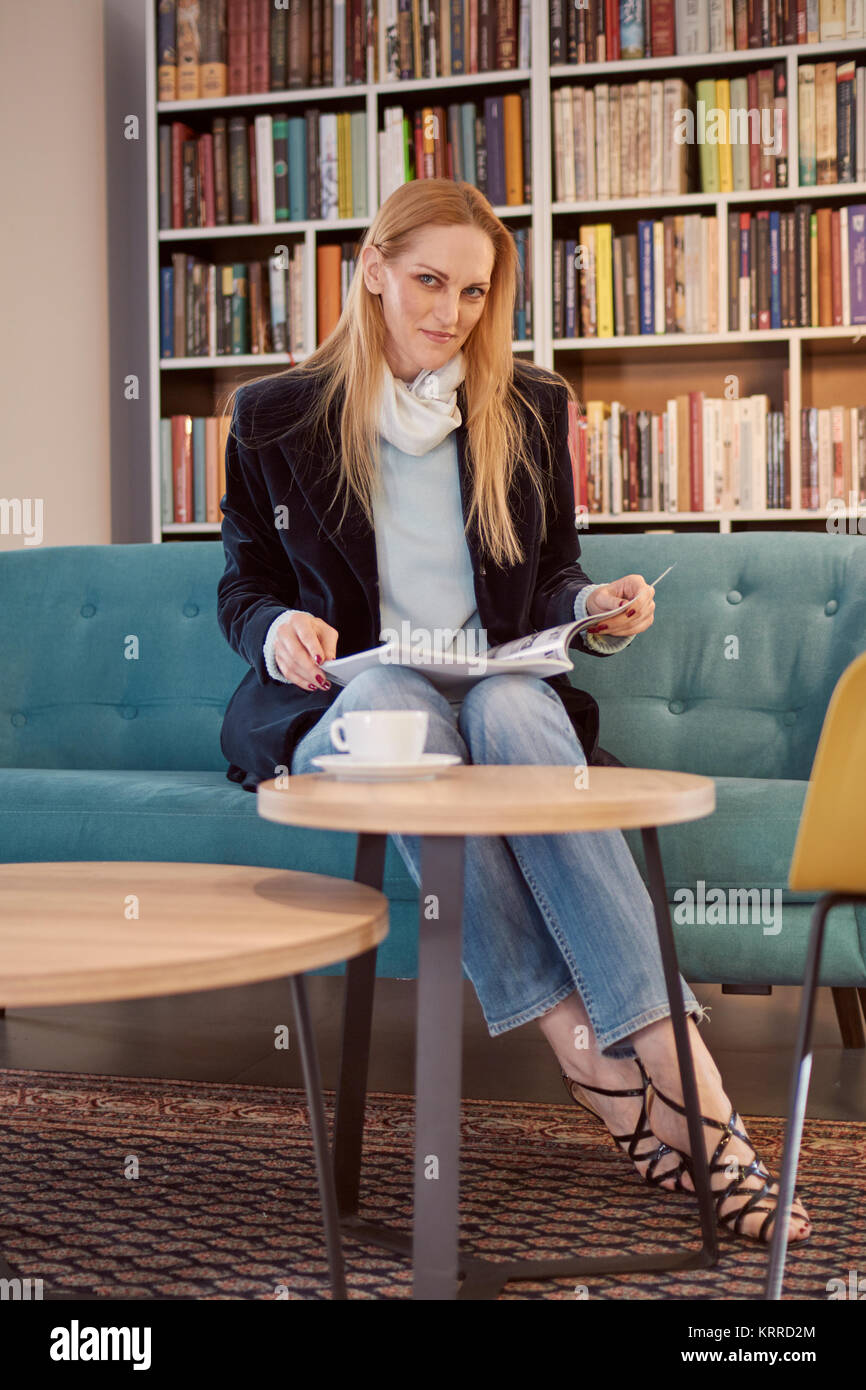 one woman sitting, 40 years old, holding magazine, book shop, book store, books on shelf behind (out of focus). Stock Photo